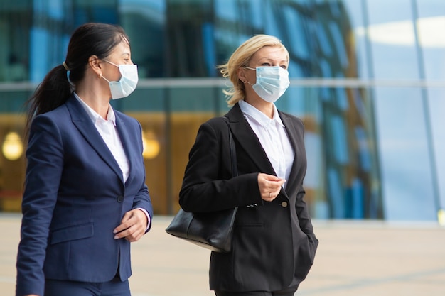 Business colleagues wearing office suits and masks, meeting and walking together in city, talking, discussing project. Medium shot. Business and coronavirus concept