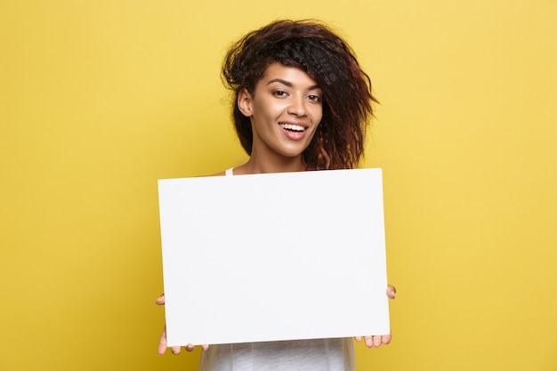 Business Concept - Close up Portrait young beautiful attractive African American smiling showing plain white blank sign. Yellow Pastel studio Background. Copy space.