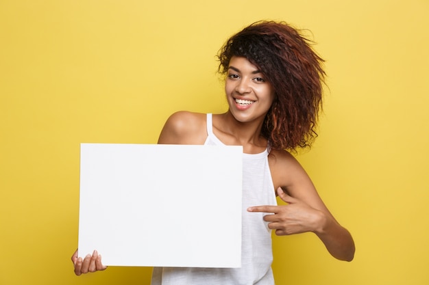 Free photo business concept - close up portrait young beautiful attractive african american smiling showing plain white blank sign. yellow pastel studio background. copy space.