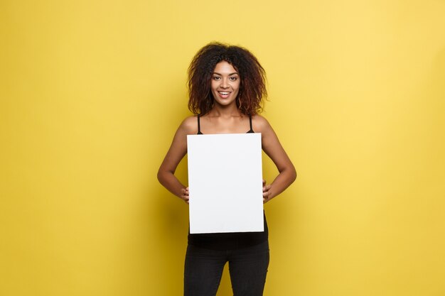 ビジネスコンセプト - クローズアップ肖像画の若い美しい魅力的なアフリカンアメリカンプレーン白い空白の看板を示す笑顔。イエローパステルスタジオの背景。スペースをコピーします。
