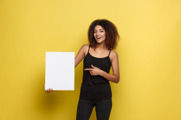 Business Concept - Close up Portrait young beautiful attractive African American smiling showing plain white blank sign. Yellow Pastel studio Background. Copy space.