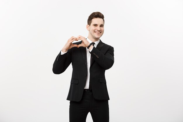 Бизнес-концепция: Портрет очаровательного привлекательного бизнесмена, держащего руки в сердечном жесте и поднимающего брови, улыбаясь, изолированного на белом сером фоне.