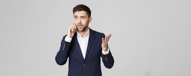 ビジネスコンセプトの肖像画若いハンサムな怒っているビジネス男スーツを着てカメラを見て電話で話している白い背景