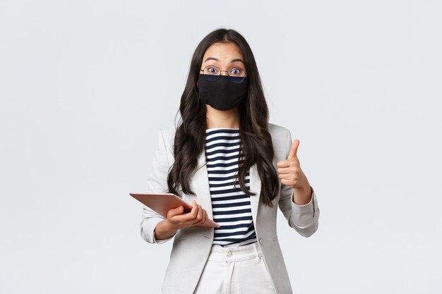 ビジネス、金融、covid-19ウイルスと社会的距離の概念を防ぎます。感銘を受けたアジアの女性オフィスマネージャー彼女の興味深いアイデア、親指を立て、保護マスクを着用し、デジタルタブレットを保持します