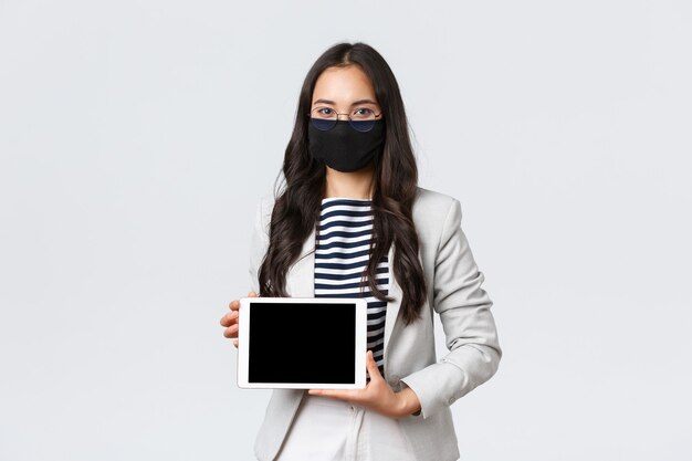 ビジネス、金融、雇用、covid-19ウイルスと社会的距離の概念を防ぎます。デジタルタブレットディスプレイとの会議でプレゼンテーションを示すアジアの女性サラリーマン、フェイスマスクを着用
