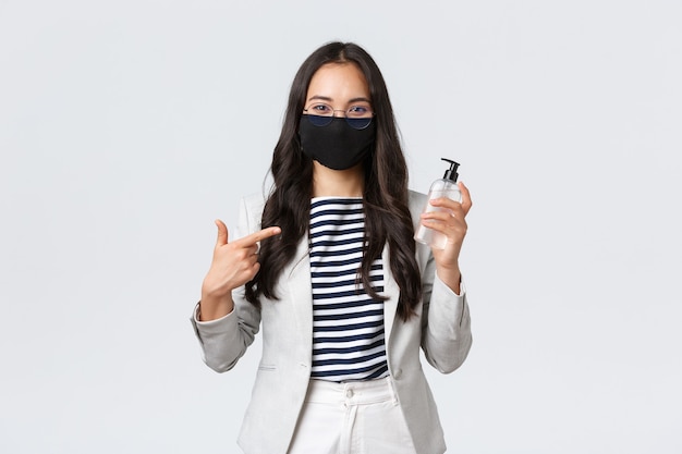 Business, finance and employment, covid-19 preventing virus and social distancing concept. Smiling cute asian office worker in face mask recommend using hand sanitizer while at work