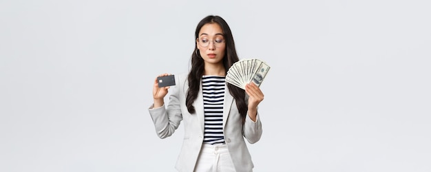 Business finance and employment entrepreneur and money concept Indecisive cute asian office lady thinking about putting cash on deposit looking curious at credit card white background