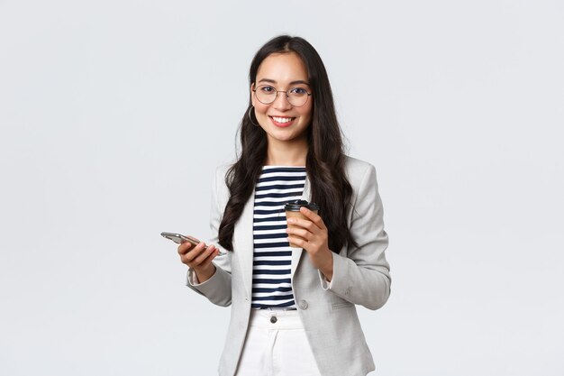 Business, finance and employment, female successful entrepreneurs concept. Professional asian businesswoman in glasses, having lunch, drinking takeaway coffee and using mobile phone