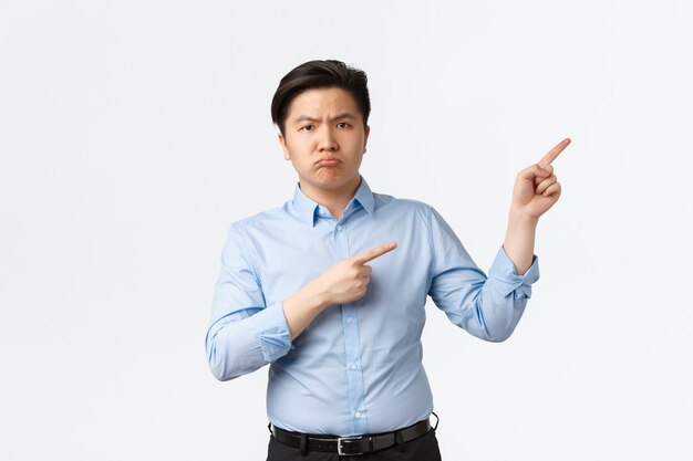 Бизнес, финансы и люди концепции. Разочарованный мрачный азиатский офисный работник, продавец в синей рубашке жалуется на что-то плохое, показывает дорогу, указывая пальцами в правом верхнем углу