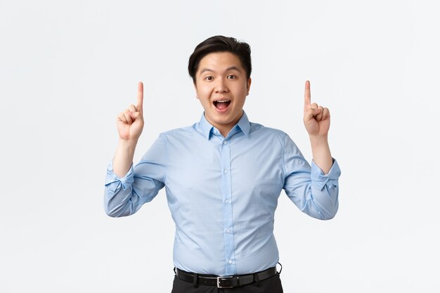 Бизнес, финансы и люди концепции. Взволнованный и изумленный азиатский бизнесмен в синей рубашке делает объявление, указывая пальцами вверх и смотрит в камеру, рассказывая важные новости, на белом фоне.