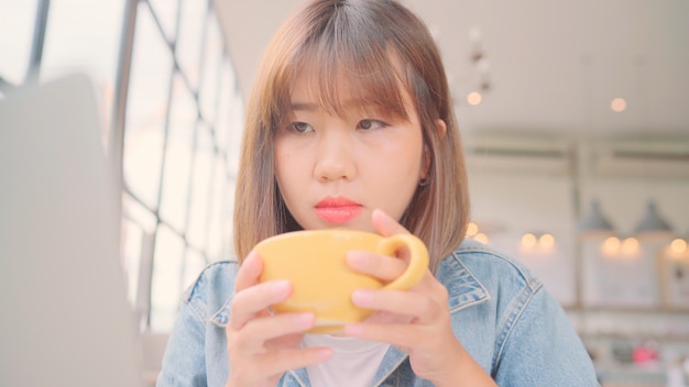 Business freelance Asian woman working, doing projects on laptop and drinking warm cup of coffee while sitting on table in cafe. 