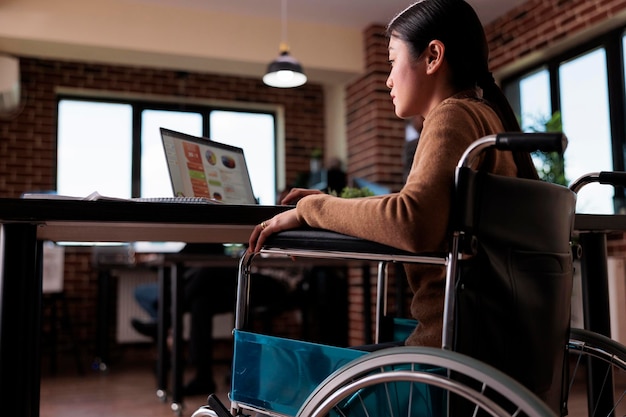 Business freelancer with physical impairment sitting in wheelchair, working on laptop in disability friendly office. Female worker with chronic health condition analyzing website presentation.