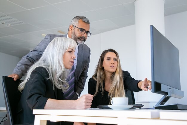 Business group watching presentation on pc monitor and discussing project, sitting at workplace with cup of coffee and pointing at display. Business communication concept