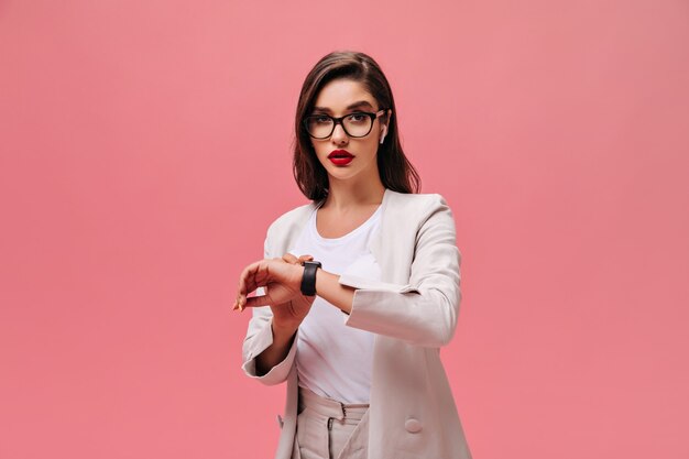 Business lady in eyeglasses looks at watch on pink background.  Beautiful serious girl with red lips in beige stylish suit posing.