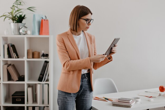 Business lady looks puzzled at tablet. Young woman in light clothing posing over office.