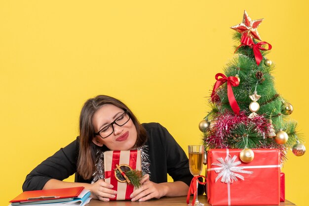 Business lady in suit with glasses holding her gift and dreaming about something sitting at a table with a xsmas tree on it in the office