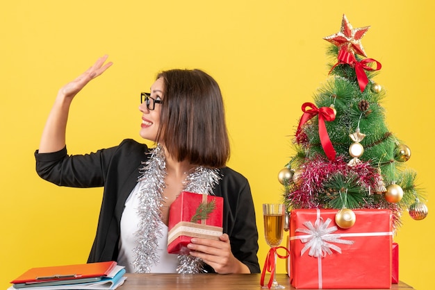 Business lady in suit with glasses holding her gift and saying bye sitting at a table with a xsmas tree on it in the office