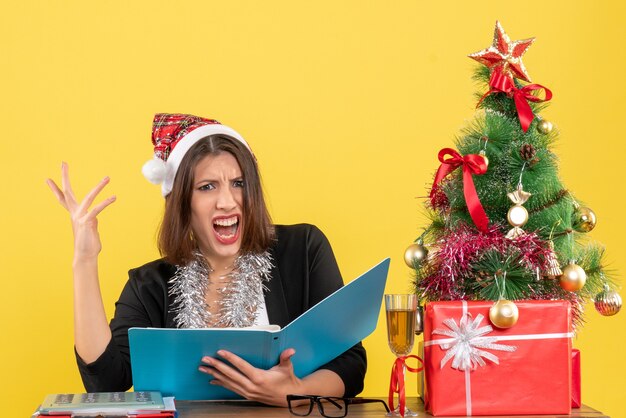 Business lady in suit with santa claus hat and new year decorations focused on document and feeling confused and sitting at a table with a xsmas tree on it in the office