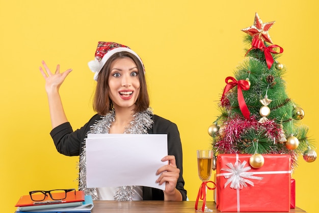 Business lady in suit with santa claus hat and new year decorations working alone pointing behind holding documents and sitting at a table with a xsmas tree on it in the office