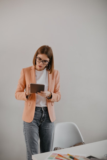 Business lady with glasses looking excitedly at tablet. Portrait of young manager in stylish outfit in white office.