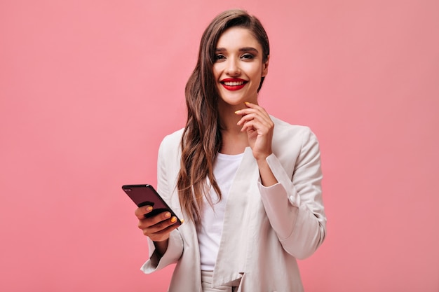 Business lady with red lips holds phone on pink background.  Curly brunette in office attire is smiling and looking at camera.