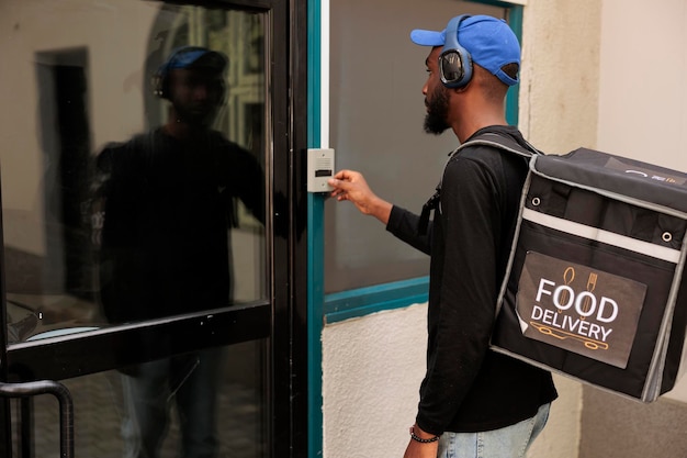 Бесплатное фото Курьер службы доставки бизнес-ланчей звонит в дверь офисного здания, ожидая клиента на улице. молодой афроамериканец с рюкзаком доставляет еду из ресторана на вынос