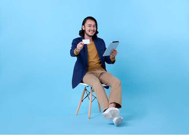 Business man asian happy smiling showing credit card and using a digital tablet while sitting on chair on bright blue.