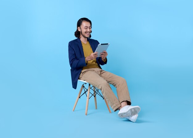 Деловой человек азиатский счастливый улыбается с помощью цифрового планшета, сидя на стуле на ярко-синем.