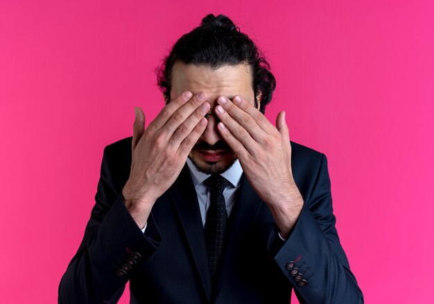 Business man in black suit and glasses covering eyes with hands standing over pink wall