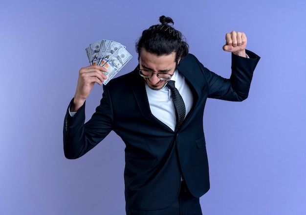 Business man in black suit and glasses holding cash happy and excited clenching fist standing over blue wall