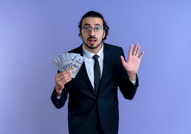 Деловой человек в черном костюме и очках, держащий деньги, смотрит вперед, смущенный поднятой рукой, стоящей над синей стеной
