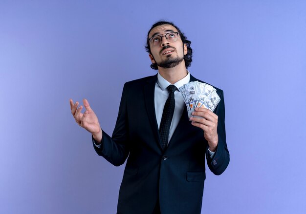 Business man in black suit and glasses holding cash looking up puzzled standing over blue wall