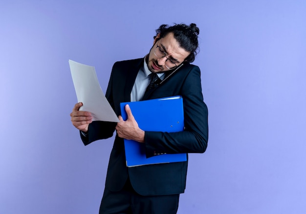Business man in black suit and glasses holding folder and documents while talking on mobile phone standing over blue wall