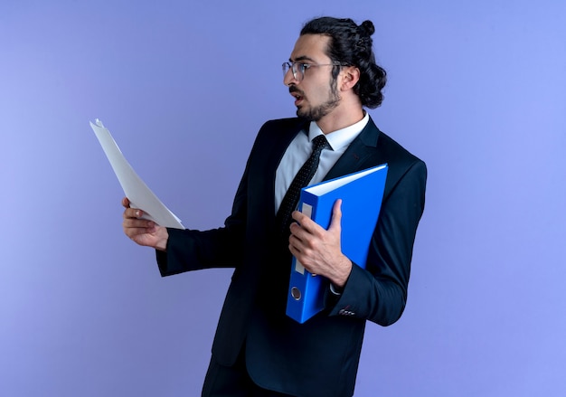 Business man in black suit and glasses holding folder looking at documents with serious face standing over blue wall