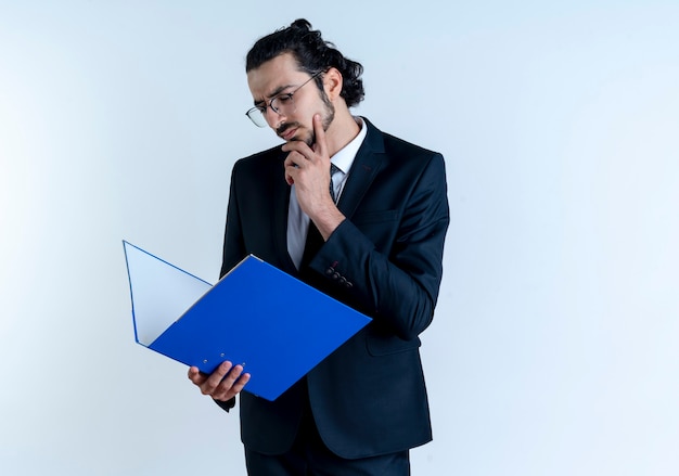 Business man in black suit and glasses holding folder, looking at it puzzled standing over white wall