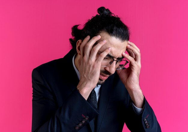 Business man in black suit and glasses holding his head with hands suffering from strong headache standing over pink wall