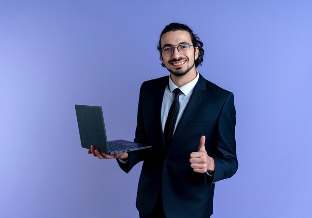 Business man in black suit and glasses holding laptop computer looking to the front smiling showing thumbs up standing over blue wall