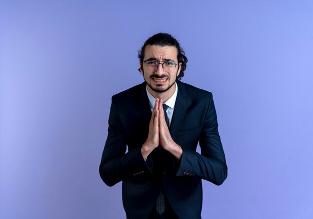 Business man in black suit and glasses holding palms together praying and begging with hope expression standing over blue wall