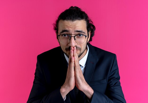 Business man in black suit and glasses holding palms together praying and begging with hope expression standing over pink wall