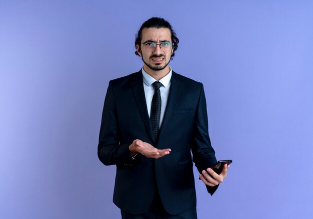 Business man in black suit and glasses holding smartphone looking to the front confused and displeased standing over blue wall