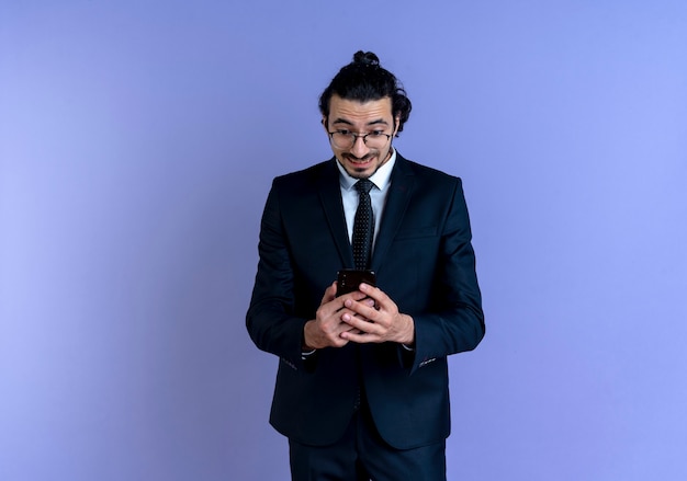 Free photo business man in black suit and glasses holding smartphone looking surprised and amazed standing over blue wall