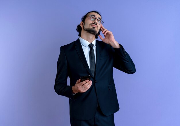 Business man in black suit and glasses holding smartphone looking up puzzled standing over blue wall