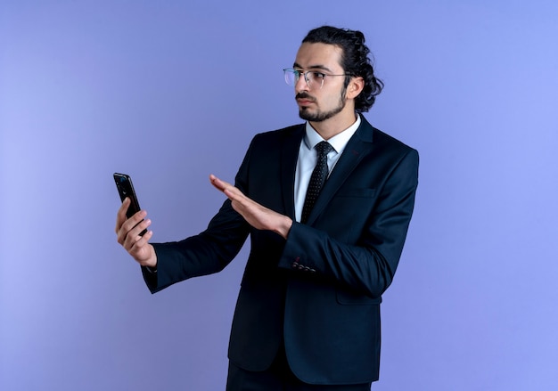 Деловой человек в черном костюме и очках держит смартфон, делая защитный жест с серьезным лицом, стоящим над синей стеной