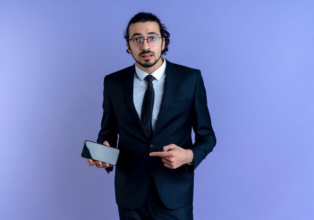 Business man in black suit and glasses holding smartphone pointing with index finger to it looking to the front confused standing over blue wall