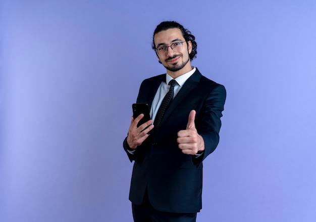 Business man in black suit and glasses holding smartphone showing thumbs up smiling confident standing over blue wall
