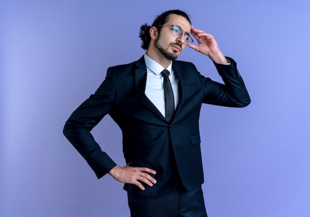 Business man in black suit and glasses looking aside confused with hand on his head standing over blue wall