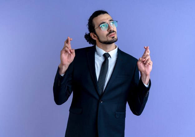 Business man in black suit and glasses looking aside making wish crossing fingers standing over blue wall