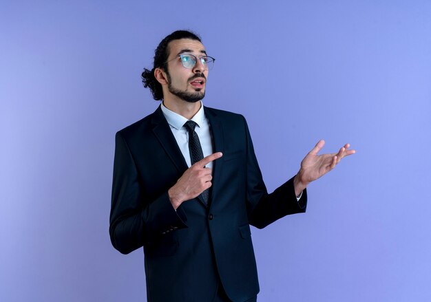 Business man in black suit and glasses looking aside pointing with index finger to the side presenting with arm of his hand standing over blue wall