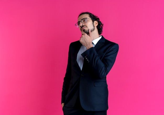 Business man in black suit and glasses looking aside with hand on chin puzzled standing over pink wall