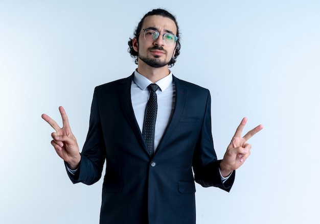 Business man in black suit and glasses looking confident showing victory sign with both hands standing over white wall